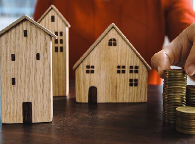 A Person Carefully Stacks Coins On A Small Wooden House Model