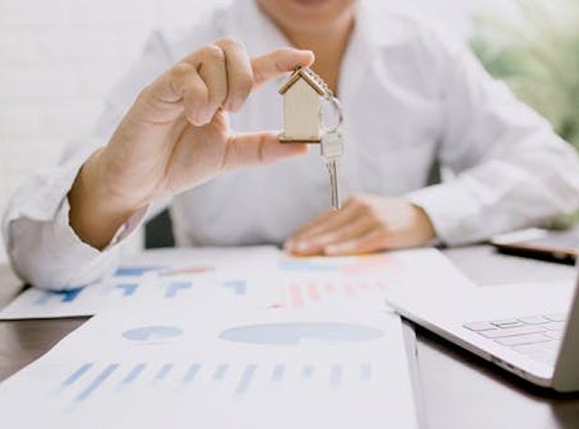 A Man Holds A House Key And A Model House In Front Of A Laptop