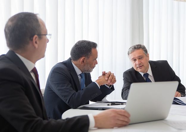 Three Business Men Seated At A Table, Each Using A Laptop, Engaged In A Discussion Or Collaboration